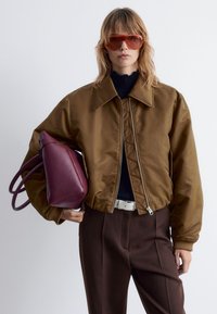 Braune Satin-Bomberjacke mit Reißverschluss, gequilteten Details und gerippten Manschetten. Model hält eine burgunderfarbene Lederhandtasche.