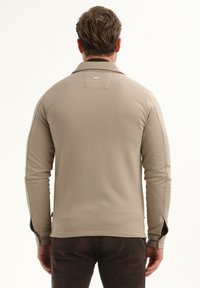 Hombre con cabello corto castaño que lleva una camisa de manga larga beige y pantalones oscuros, visto desde atrás contra un fondo blanco liso.