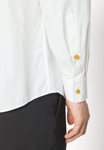 White cotton shirt featuring a tailored fit, pointed cuff, and contrasting yellow buttons. The shirt is tucked into dark trousers.