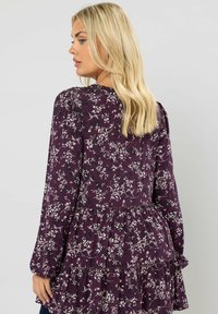 Lila kukallinen paita pitkähihaisilla hihoilla, kerrostetuilla olkahapsuilla ja kerrostetulla röyhelöhelmalla. Kankaan pinta on pehmeä ja siinä on kasvitieteellinen kuvio.