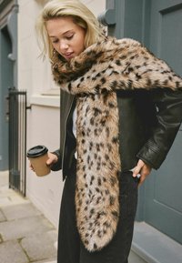 Écharpe en fausse fourrure avec un imprimé léopard tan et noir, offrant une texture douce et une forme longue et étroite, portée sur une veste en cuir noire.