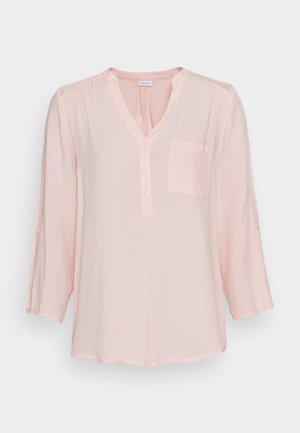 Lichtroze blouse van zachte stof, met een V-hals, knoopsluiting en een enkele borstzak. Driekwart mouwen met oprol-tabs.