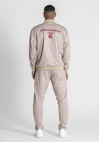 Beige trainingspak met herhalend logopatroon, bestaande uit een zip-up jas en broek met elastische tailleband. Gecamoufleerd met ribboorden aan de mouwen en de kraag.