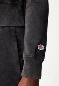 Črna majica s kapuco z rebrastimi manšetami, mehko teksturo in izstopajočim vezenim logotipom v modri in rdeči barvi na desnem rokavu.