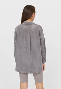 Grå manchester-skjortjacka med krage, långa ärmar och en veckdetalj på baksidan. Slät textur och avslappnad passform.