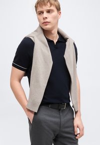 Νέος άνδρας που φοράει μια μαύρη πόλο μπλούζα και γκρι παντελόνι με ένα ανοιχτό μπεζ πουλόβερ περασμένο πάνω από τους ώμους του, κοιτάζοντας προς τα πλάγια.