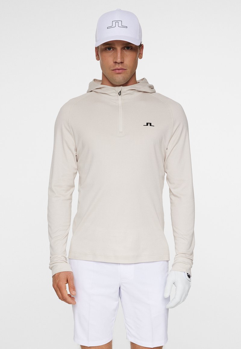 Beige långärmad hoodie med dragkedja, med en svart logotyp på bröstet. Matchad med vita shorts och en vit keps med svarta detaljer.