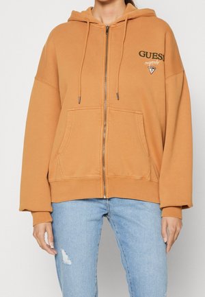 Person iført en lysebrun lynlåshoodie med Guess-logo på brystet og lyseblå slidte jeans, stående mod en ensfarvet baggrund.