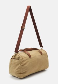 Kangas duffel-laukku vaaleassa beige-värissä, jossa on ruskeat nahkaosuudet ja säädettävä hihna. Pyöreä muoto, jossa on kaksinkertaiset kantokahvat ja vetoketjukiinnitys.