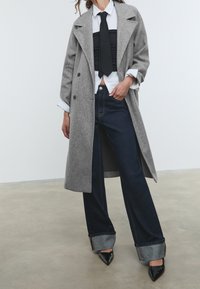 Grijze wollen overjas, witte shirt, zwarte stropdas en donkerde brede jeans met omgeslagen pijpen. Zwarte puntige hakken. Gladde texturen en een getailleerde pasvorm.