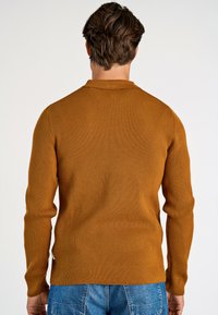 Pull à manches longues côtelé dans une teinte de brun chaud avec une texture subtile. Doté d'un col classique et d'un détail de tag sur le côté.