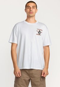 T-shirt en coton blanc avec un graphique sur le devant, agrémenté d'accents marron et noirs. Manches courtes, col rond et coupe décontractée.