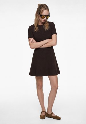 Kobieta o długich blond włosach, ubrana w czarną sukienkę z krótkim rękawem, żółte okulary przeciwsłoneczne i brązowe płaskie buty, stojąca z założonymi ramionami.