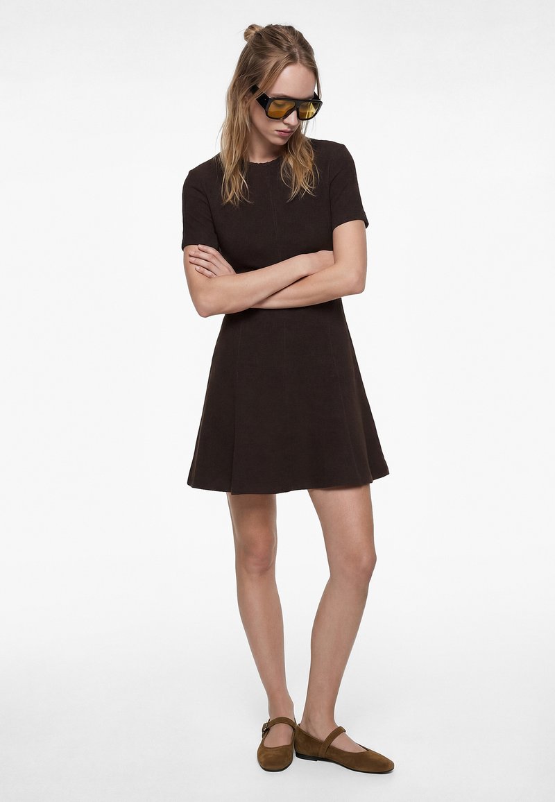 Kobieta o długich blond włosach, ubrana w czarną sukienkę z krótkim rękawem, żółte okulary przeciwsłoneczne i brązowe płaskie buty, stojąca z założonymi ramionami.