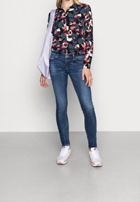 Kvinna i blommig långärmad skjorta, blå skinny jeans, vitt-rosa sneakers och bär en ljuslila axelväska, står på en grå golv.