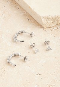 Silberne Creolen- und Steckerohrringe mit einem strukturierten Design aus kleinen Perlen und klaren Edelsteinen, die auf einer hellen Steinoberfläche liegen.