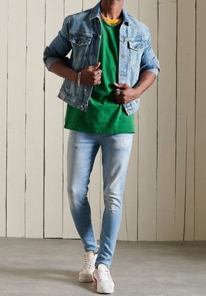 Veste en denim clair, t-shirt vert, jeans slim bleu clair et baskets blanches avec des rayures rouges. Les accessoires comprennent une chaîne en or et des bracelets.