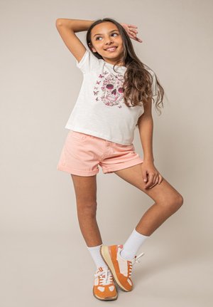 Fille souriante aux longs cheveux posant, portant un t-shirt blanc avec un motif papillon et crâne, un short pêche, des chaussettes blanches et des baskets orange et blanches.