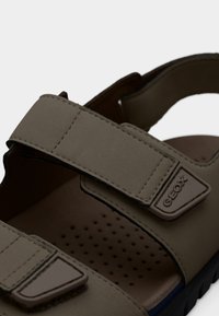 Καφέ συνθετικά σανδάλια με δύο ρυθμιζόμενους ιμάντες, διάτρητη σόλα και λογότυπο Geox στο πλάι. Επιφάνεια με υφή και κυρτές σόλες.