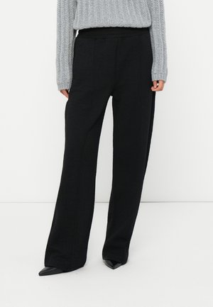Woman wearing black textured wide-leg trousers, gray knitted sweater, and black pointed-toe shoes standing against a white background.