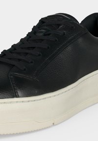 Schwarzer Ledersneaker mit schwarzen Schnürsenkeln und dicker weißer Gummisohle, zeigt obere und seitliche Details vor einfachem Hintergrund.
