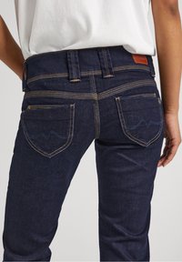 Mörkblå jeans av denim med insvängd midja, synliga sömmar, två bakfickor och en läderlogotyp på midjebandet.