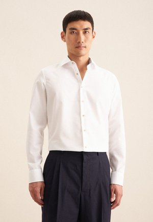 Weißes Langarmhemd mit geknöpftem Kragen und hellbeigen Knöpfen, kombiniert mit marineblauen, gefalteten Hosen. Glattes Baumwollmaterial, taillierte Passform.