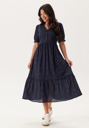 Marineblauwe midi-jurk met puffmouwen, knopen aan de voorkant, bloemig eyeletpatroon en gelaagde rok; lichtgewicht stof en vloeiende silhouet.