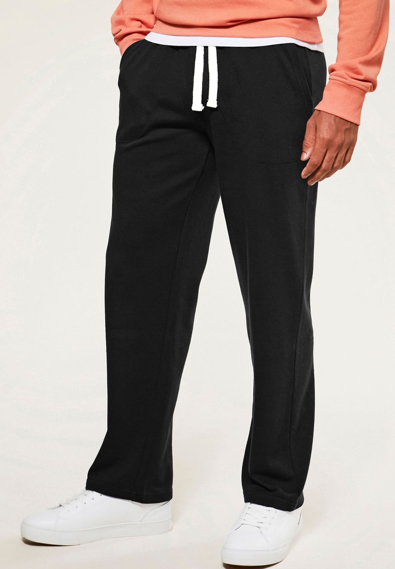 Man wearing casual black drawstring pants paired with white sneakers and a coral long-sleeve top, standing against a plain background.