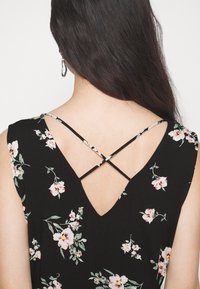 Femme aux longs cheveux foncés portant une robe noire fleurie avec de fines bretelles croisées dans le dos ouvert et des boucles d'oreilles créoles argentées.