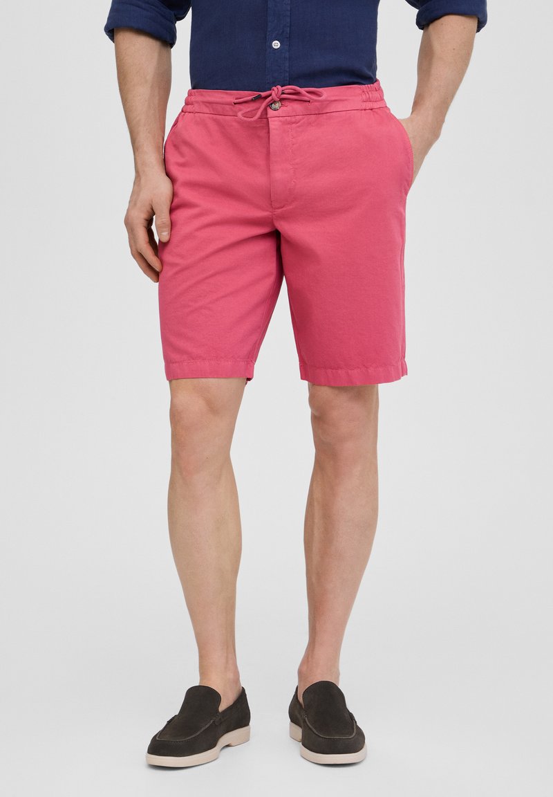 Shorts en coton rose avec une taille élastique et un cordon de serrage. Comprend des poches latérales et une coupe simple, associés à des chaussures slip-on sombres.