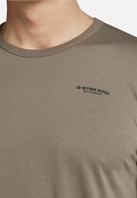 Zbliżenie osoby w beżowej koszulce z okrągłym dekoltem, z małym czarnym napisem "G-STAR RAW" na lewej piersi.