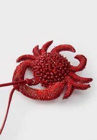Colgante en forma de cangrejo con cuentas rojas, con un cuerpo y patas texturizadas, atado a un cordón rojo, sobre un fondo blanco liso.