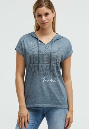 Junge Frau trägt ein blaues kurzärmeliges Kapuzenshirt mit dem Text „Salut Joie de Vivre Joy of Life“ und blaue Jeans, steht vor schlichtem Hintergrund.