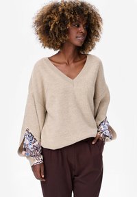 Beżowy sweter dzianinowy z dekoltem w szpic i szerokimi rękawami, z niebiesko-brązowym wzorzystym akcentem na mankietach, w zestawieniu z brązowymi spodniami.