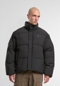 Homme aux cheveux blonds courts portant une veste noire rembourrée et un pantalon cargo marron, regardant sur le côté sur un fond blanc.
