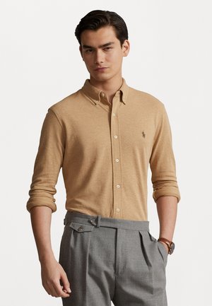 Camisa de botonadura ligera de color marrón con cuello y mangas enrolladas, combinada con pantalones grises con pliegues. Presenta un pequeño emblema en el pecho.