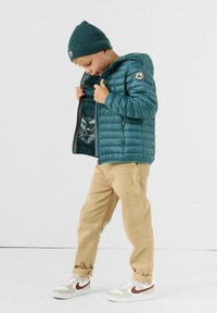 Turkooise gewatteerde jas met capuchon, voorzien van een ritssluiting en geribbelde manchetten, gedragen over een grafisch T-shirt. Beige broek en witte sneakers met rode accenten.