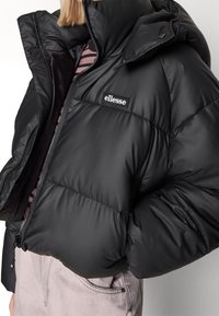 Czarna pikowana kurtka puchowa z wysokim kołnierzem, zamkiem błyskawicznym i naszywką z logo. Posiada przeszywane sekcje dla izolacji i gładkie wykończenie.