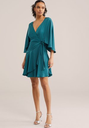 Nő, aki türkiz színű, széles ujjú wrap ruhát visel ezüst magassarkú szandálokkal, világos, egyszínű háttér előtt áll.