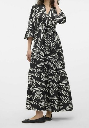 Kobieta ubrana w czarno-białą sukienkę kopertową w kwiatowy wzór z rękawami do połowy łokcia i czarne baleriny, stojąca na jednolitym tle.