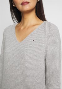 Pull gris clair en maille avec col en V et manches longues. Présente un petit logo sur la poitrine et un tissu texturé pour un intérêt visuel.