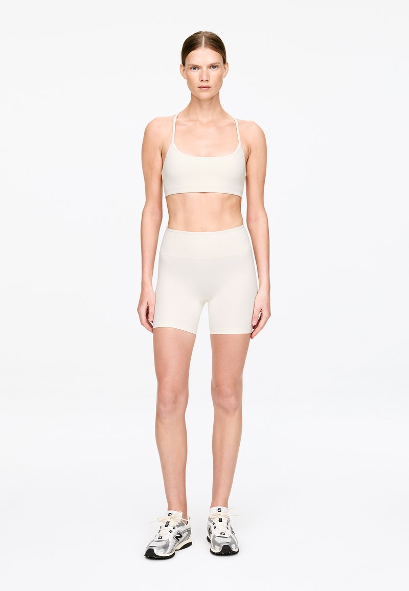 Ensemble de sport beige clair composé d'un haut ajusté et d'un short, avec une texture lisse. Porte avec des baskets argentées, style minimaliste.