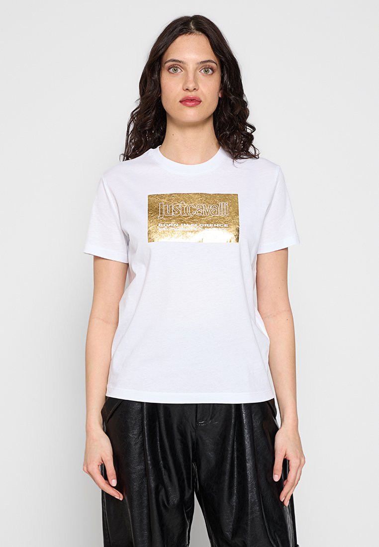 Camiseta blanca de algodón con un estampado de logotipo rectangular metálico en dorado, cuello redondo y mangas cortas. Combinada con pantalones sueltos negros.