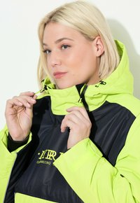 Kobieta ściągająca sznurek przy kołnierzu neonowo żółtej i czarnej kurtki z kapturem.