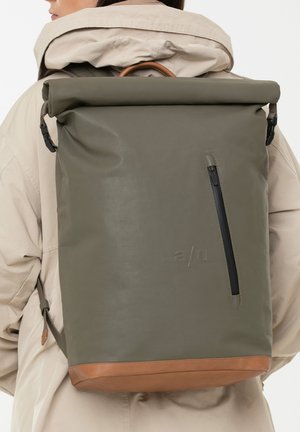 Person trägt eine beige Jacke und einen hohen, olivgrünen Rolltop-Rucksack mit vertikalem Reißverschluss und braunem Lederboden.