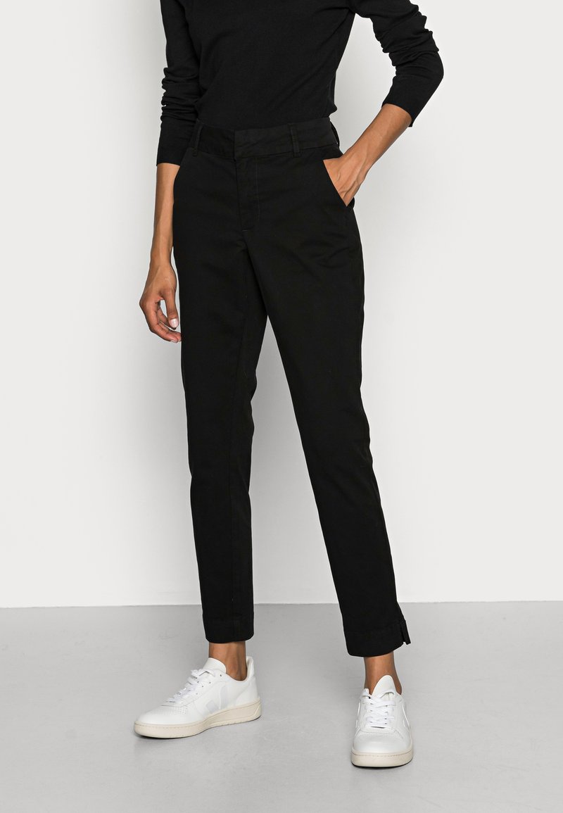 Pantalons noirs fuselés avec une texture lisse et un léger éclat, dotés de poches latérales et d'une coupe slim, associés à des baskets blanches.