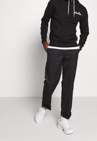 Sudadera negra con bolsillo frontal y cordones blancos, combinada con pantalones negros holgados que tienen una cinturilla blanca y puños enrollados. Zapatillas blancas.