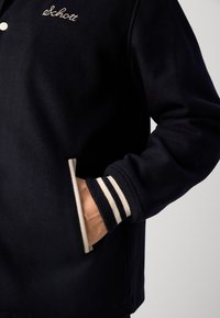 Tumši zila jaka ar ribotām piedurknēm, kurām ir baltas svītras, ar pielāgotu piegriezumu un sānu kabatu. "Schott" ir izšūts zelta krāsā.