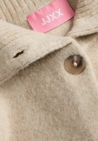 Cappotto di lana beige con un bottone marrone grigiastro e un'etichetta di stoffa rosa con la scritta "JJXX" cucita all'interno del colletto.